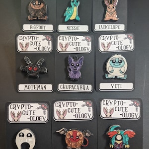 May include: Nine enamel pins featuring various cryptids, including Bigfoot, Nessie, Jackalope, Mothman, Chupacabra, Yeti, Nightcrawler, Jersey Devil, and Snallygaster. Each pin has a black background with white text that reads "Crypto-Cute-ology".