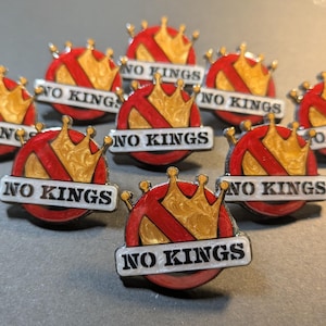 Może przedstawiać: Kolekcja emaliowanych przypinek z czerwono-złotym motywem korony i napisem "NO KINGS" na białym banerze. Przypinki mają okrągły kształt, srebrną obwódkę i błyszczące wykończenie.