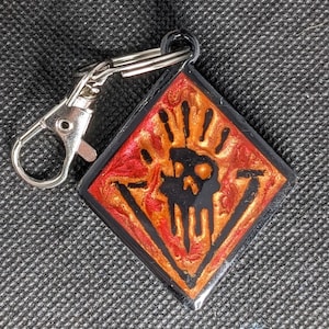 Peut inclure: Un porte-clés en forme de diamant noir et rouge avec un motif de crâne et de main. Le porte-clés a un anneau en métal argenté.