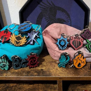 May include: A collection of colorful keychains with various emblem designs. The keychains feature a variety of shapes and colors, including blue, red, gold, and green. Each keychain has a metal ring for attaching to keys or bags.