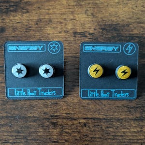 Puede incluir: Dos pares de pendientes de bot&oacute;n en tarjetas negras. Un par presenta un dise&ntilde;o plateado de estrella, el otro un rayo amarillo. Las tarjetas dicen "ENERGY" y "Little Root Traders."