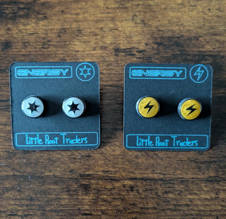 Puede incluir: Dos pares de pendientes de bot&oacute;n en tarjetas negras. Un par presenta un dise&ntilde;o plateado de estrella, el otro un rayo amarillo. Las tarjetas dicen "ENERGY" y "Little Root Traders."