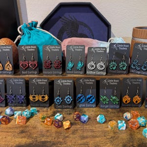 Può includere: Esposizione di orecchini tagliati al laser con vari design, tra cui loghi D&D e simboli di classi fantasy. Gli orecchini sono su cartoncini neri con la scritta "Little Root Traders". Dadi colorati sono sparsi su una superficie di legno.
