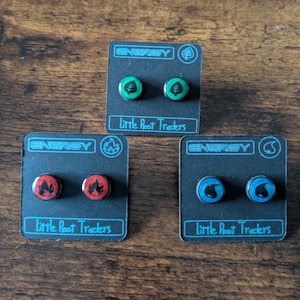 Puede incluir: Tres pares de pendientes de bot&oacute;n en tarjetas negras. Cada par presenta un dise&ntilde;o diferente: una hoja verde, una llama roja y una gota de agua azul. La palabra "ENERGY" est&aacute; impresa en la parte superior de cada tarjeta, con "Little Root Traders" debajo.