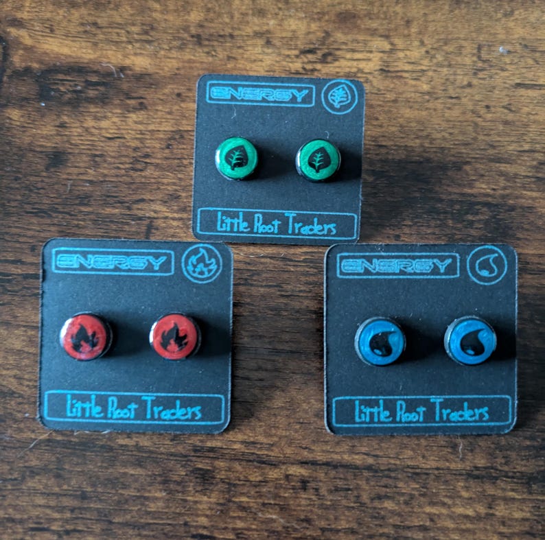 Puede incluir: Tres pares de pendientes de bot&oacute;n en tarjetas negras. Cada par presenta un dise&ntilde;o diferente: una hoja verde, una llama roja y una gota de agua azul. La palabra "ENERGY" est&aacute; impresa en la parte superior de cada tarjeta, con "Little Root Traders" debajo.