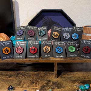 May include: A selection of enamel pins on black cards, each displaying a fantasy class symbol. The hexagonal pins have a metallic border. The cards are labelled with class names like "Druid Pin" and "Sorcerer Pin". The pins are arranged on a wooden surface.