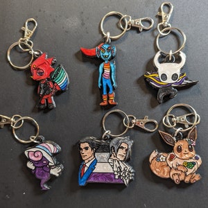 May include: A set of ten colorful keychains featuring various characters. The keychains are made of painted metal and feature a variety of designs, including a blue and red character with a crown, a black and white character with a cape, and a brown and white character with a flower crown.