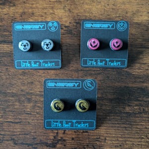 Puede incluir: Tres pares de pendientes de bot&oacute;n en tarjetas negras. Los pendientes tienen un dise&ntilde;o circular con la palabra "ENERGY" encima. Los pendientes son plateados, rosas y dorados. El texto "Little Root Traders" est&aacute; debajo.