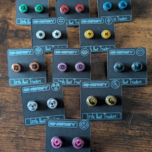 May include: A collection of stud earrings with various designs, including lightning bolts, stars, and flames. Each pair is mounted on a black card with the word "ENERGY" and "Little Root Traders" printed on it. The earrings are in various colors.