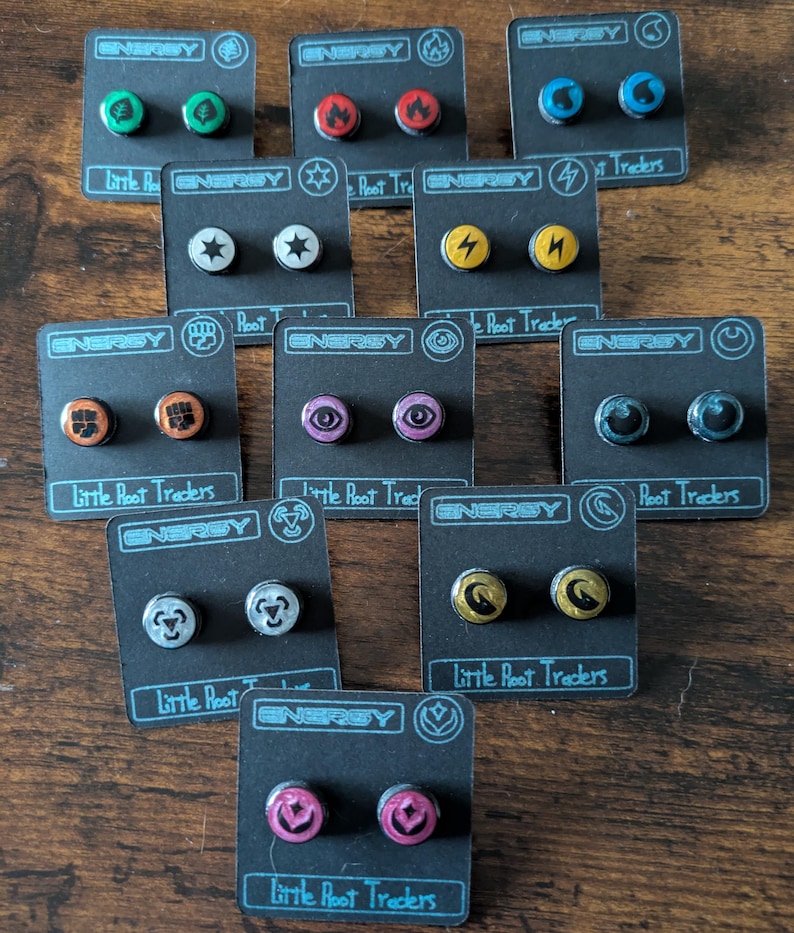 Puede incluir: Una colecci&oacute;n de pendientes de bot&oacute;n con varios dise&ntilde;os, incluyendo rayos, estrellas y llamas. Cada par est&aacute; montado en una tarjeta negra con la palabra "ENERGY" y "Little Root Traders". Los pendientes son de varios colores.