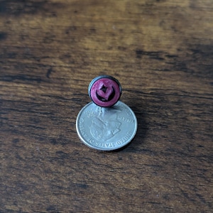 Puede incluir: Un peque&ntilde;o pendiente de bot&oacute;n redondo de color rosa oscuro con un dise&ntilde;o en forma de coraz&oacute;n, colocado encima de una moneda de veinticinco centavos estadounidense. El pendiente tiene un borde negro.