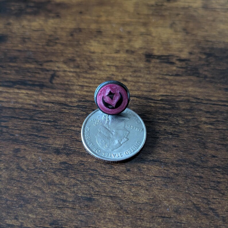 Puede incluir: Un peque&ntilde;o pendiente de bot&oacute;n redondo de color rosa oscuro con un dise&ntilde;o en forma de coraz&oacute;n, colocado encima de una moneda de veinticinco centavos estadounidense. El pendiente tiene un borde negro.