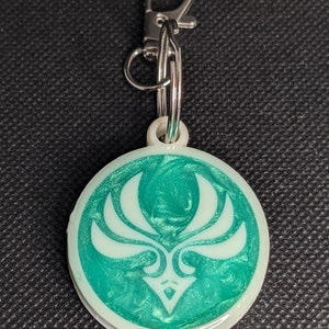 May include: A round, green and white keychain with a stylized, white design. The keychain has a silver metal ring and a silver metal clasp.