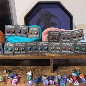 May include: A display of stud earrings with various designs, including Warlock, Rogue, Sorcerer, Druid, Monk, and more. Each pair is mounted on a black card with the "Little Root Traders" logo. A collection of colorful dice is in the foreground.