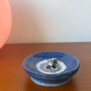 Puede incluir: Un pequeño plato redondo de cerámica con un esmalte azul oscuro y un interior blanco y azul claro. Dos anillos de plata descansan dentro del plato. El plato está sobre una superficie de madera.