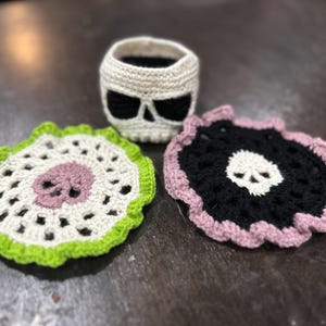 May include: Three crocheted skull-shaped coasters. One is white with a black skull, one is black with a white skull, and one is white with a pink skull and a green border.
