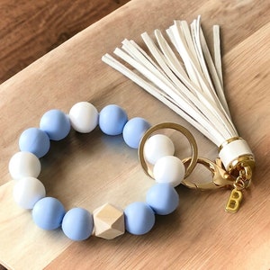May include: A light blue and white beaded keychain with a wooden bead and a white tassel. The keychain is attached to a gold key ring.