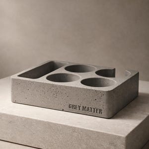 Puede incluir: Un organizador de escritorio rectangular de hormigón gris con una textura lisa y moteada, que presenta cuatro aberturas circulares y una ranura rectangular alargada. Las palabras GREY MATTER están grabadas en el frente de la estructura.