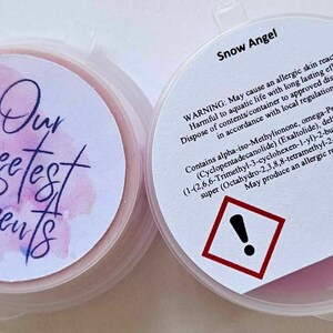 May include: Two round, clear plastic containers with lids. The container on the left has a pink and white label with the text "Our Sweetest Scents". The container on the right has a white label with the text "Snow Angel" and a warning label with an exclamation point.
