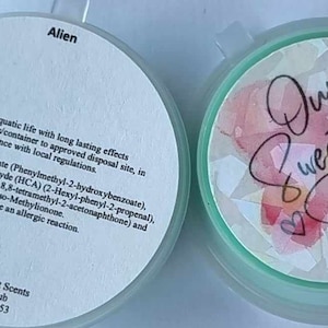 May include: A round, white container with a green lid. The container has a label that reads "Alien" and includes a list of ingredients. The lid has a colourful design with the text "Our Sweetest Scents".