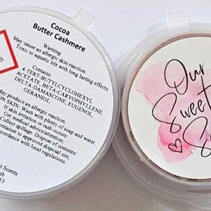 May include: A white plastic container with a red warning label and text that reads "Cocoa Butter Cashmere" and includes a list of ingredients. The container is labelled "Our Sweetest Scents" with a phone number and address. The container is sitting on a white surface.