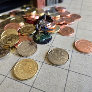 Puede incluir: Un conjunto de monedas doradas, plateadas y cobrizas con dise&ntilde;os detallados. Una figura en miniatura de un guerrero se encuentra entre las monedas. Las monedas est&aacute;n dispuestas sobre una superficie cuadriculada, sugiriendo un juego de mesa.