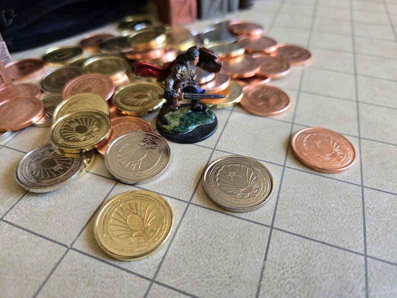 Puede incluir: Un conjunto de monedas doradas, plateadas y cobrizas con dise&ntilde;os detallados. Una figura en miniatura de un guerrero se encuentra entre las monedas. Las monedas est&aacute;n dispuestas sobre una superficie cuadriculada, sugiriendo un juego de mesa.