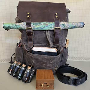 Może przedstawiać: Szary plecak z woskowanego płótna z brązowymi skórzanymi detalami, zawierający zwiniętą mapę, książki i fiolki. Zawiera drewniane pudełko, czarny pasek i skórzany uchwyt na fiolki ze szklanymi butelkami. Mapa głosi "Crystal River" i "Highpass".