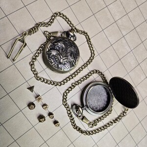 May include: A silver pocket watch with a detailed dragon design on the cover, attached to a chain. Beside it are several dice, including a D20, D12, D8, D6, and D4, all in a bronze color. The watch is open, revealing the inside.