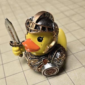 Op de afbeelding: Een geel rubberen eendje verkleed als middeleeuwse ridder. De eend draagt een zwarte en bronzen helm en bijpassend harnas met zilveren accenten. Het houdt een klein zilveren zwaard in de rechtervleugel en heeft een rond schild aan de zijkant.