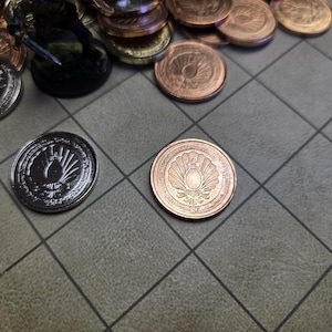 Puede incluir: Primer plano de varias monedas met&aacute;licas con dise&ntilde;os detallados, algunas plateadas y otras de color cobre. Las monedas est&aacute;n dispersas sobre una superficie gris con patr&oacute;n de cuadr&iacute;cula, con una figura en miniatura en el fondo.