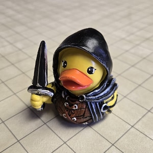 Könnte beinhalten: Eine gelbe Gummiente mit schwarzem Kapuzenmantel und schwarzem Dolch. Die Ente hat einen orangefarbenen Schnabel und trägt ein braunes und schwarzes Outfit. Es ist ein Neuheitenartikel.