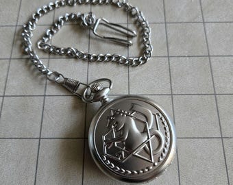 May include: A silver-toned pocket watch with a circular case featuring an embossed dragon design inside a geometric star shape. The watch is attached to a matching silver-colored metal chain that includes a small hook clasp for secure attachment.