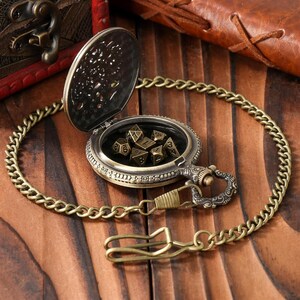 May include: An antique brass pocket watch with a hinged lid, revealing a set of polyhedral dice. The watch is attached to a matching chain with a clasp. The watch and chain are on a wooden surface.