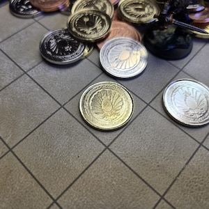 Puede incluir: Una colecci&oacute;n de monedas met&aacute;licas con dise&ntilde;os en relieve detallados. Las monedas son de color plata, oro y cobre. Algunas monedas est&aacute;n dispersas sobre una superficie con un patr&oacute;n de cuadr&iacute;cula, lo que sugiere un juego o un objeto de colecci&oacute;n.