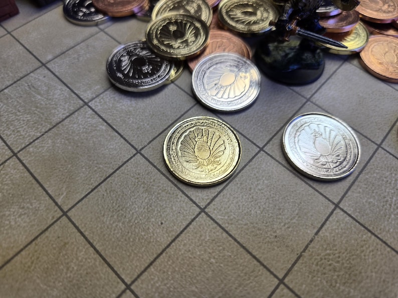 Puede incluir: Una colecci&oacute;n de monedas met&aacute;licas con dise&ntilde;os en relieve detallados. Las monedas son de color plata, oro y cobre. Algunas monedas est&aacute;n dispersas sobre una superficie con un patr&oacute;n de cuadr&iacute;cula, lo que sugiere un juego o un objeto de colecci&oacute;n.