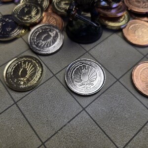 Puede incluir: Varias monedas met&aacute;licas en tonos dorados, plateados y bronce, dispuestas sobre una superficie gris con patr&oacute;n de cuadr&iacute;cula. Las monedas presentan un dise&ntilde;o de emblema detallado, posiblemente para un juego de fantas&iacute;a.