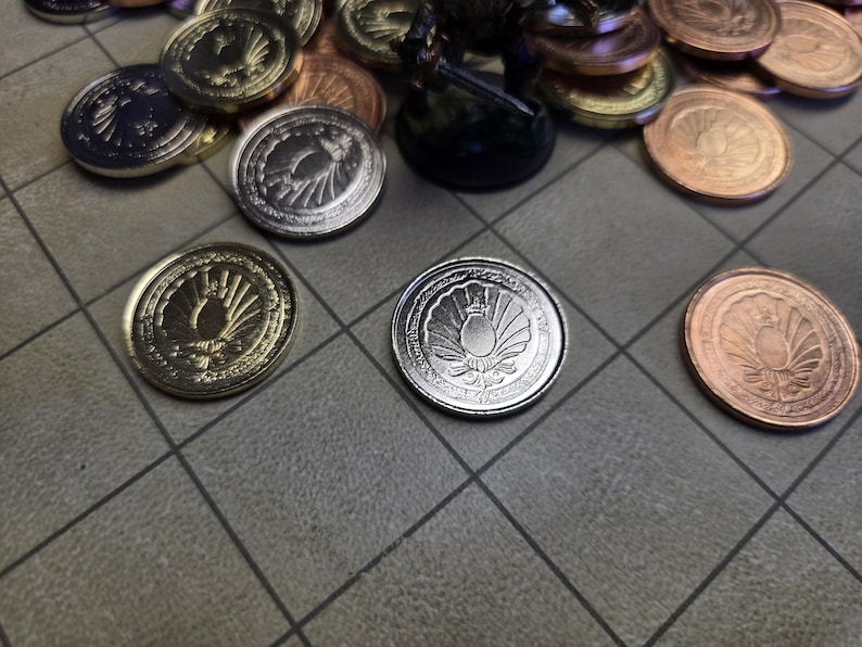 Puede incluir: Varias monedas met&aacute;licas en tonos dorados, plateados y bronce, dispuestas sobre una superficie gris con patr&oacute;n de cuadr&iacute;cula. Las monedas presentan un dise&ntilde;o de emblema detallado, posiblemente para un juego de fantas&iacute;a.