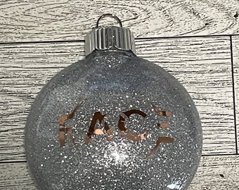 Op de afbeelding: Een zilveren glitter kerstbal met het woord "FACE" in gouden glitter.