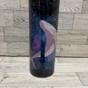Könnte beinhalten: Ein Edelstahlbecher mit einem lila Wal-Design vor einem dunkelblauen und schwarzen Galaxiehintergrund. Der Becher hat einen transparenten Deckel und ist für heiße oder kalte Getränke konzipiert. Ein stilvolles und funktionales Trinkgefäß.