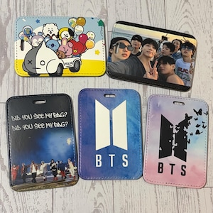 May include: Five rectangular luggage tags with various designs. One tag features a cartoon with balloons and a van. Another displays a group of people. The remaining tags have the BTS logo, one in white on blue, and another with butterflies on pink.