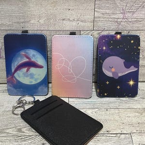 Pode incluir: Uma coleção de porta-cartões com designs únicos. Um apresenta uma baleia saltando em frente a uma lua cheia, outro tem um design em forma de coração e o terceiro mostra uma baleia em um céu estrelado. Um porta-cartões preto com chaveiro também está incluído.