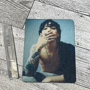 Puede incluir: Un ambientador rectangular con una persona tatuada con una parte superior negra. Una botella de spray transparente está al lado del ambientador. El ambientador tiene un fondo azul claro.