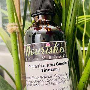 Puede incluir: Una botella de vidrio marrón con un gotero y una etiqueta blanca que dice "Nourished by Embrace" y "Parasite and Candida Tincture". La etiqueta enumera los ingredientes, incluyendo nuez negra, clavo, raíz de uva de Oregón y alcohol (45%). La botella es de 2 oz y está etiquetada para tomar 2-3 gotas debajo de la lengua, 8-10 oz de bebida.