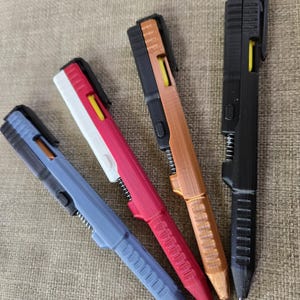 May include: Four pens in various colors: blue, red, copper, and black. Each pen has a black top, a yellow accent, and a silver spring. The pens are lying on a textured, tan surface.
