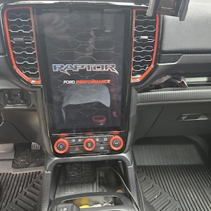 May include: A vehicle dashboard featuring a large vertical touchscreen display with the text RAPTOR FORD PERFORMANCE. The console includes black air vents with orange trim, circular climate control knobs with orange rings, and a gear shifter.