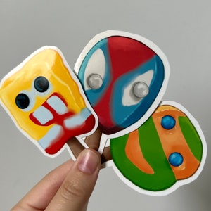Könnte beinhalten: Drei bunte Popsicle-Aufkleber mit einem gelben Schwamm-Charakter, einem roten und blauen Superhelden und einem grünen und orangefarbenen Schildkröten-Charakter.
