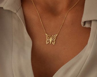 May include: A gold-colored butterfly pendant necklace. The butterfly design is intricately cut out, and the necklace is worn on a person's neck. The background is a neutral color.