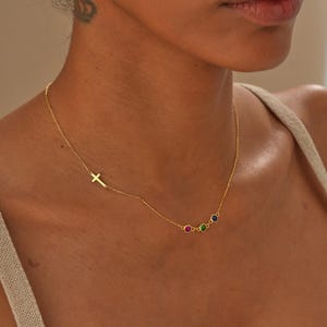 May include: A delicate gold necklace featuring a small cross pendant and three colorful gemstone accents. The necklace is worn on a person's neck, showcasing its elegant design and vibrant gemstones.