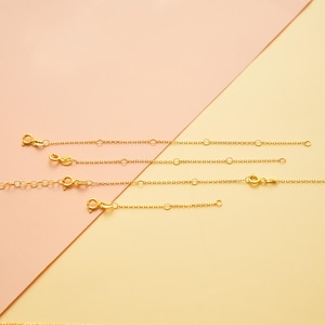 May include: Gold-colored necklace extenders with lobster claw clasps and small chain links are displayed against a two-tone pink and yellow background. The extenders vary in length, offering options for adjusting necklace sizes.